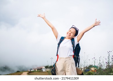 Young Smiling Asian Woman With Backpack Enjoying Travel. Beautiful Tourist Female Raised Hands With Background Of Fog And Village On Mountain. Freedom And Happy Life Concept.