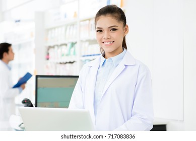 Young Smiling Asian Woman Pharmacist In White Gown Coat Uniform Working On Laptop Computer In Chemist Shop Or Pharmacy