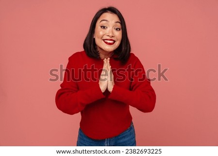 Young smiling asian woman in red sweater doing pray gesture with folded hands isolated over pink background