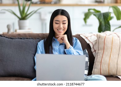 Junge lächelnde asiatische Frau, die auf dem Sofa sitzt und Laptop benutzt, um soziale Netzwerke zu durchsuchen, einkaufen. Gutes Zuhause