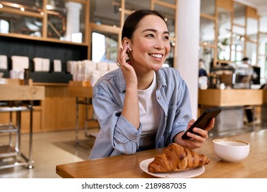 Young smiling Asian woman student using smartphone for elearning wearing earbud, watching online webinar, listening podcast music audio content streaming in mobile app on cell phone sitting in cafe.