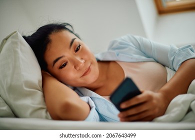 Young Smiling Asian Woman Texting On Mobile Phone While Lying Down On Bed At Home.