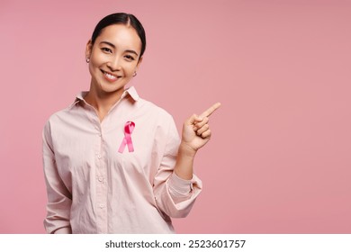 Young, smiling Asian woman wearing pink ribbon looking a camera showing, pointing finger on pink background. Breast cancer awareness concept - Powered by Shutterstock