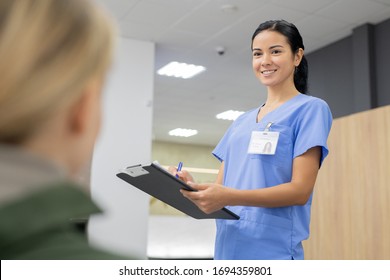 Young smiling assistant in blue uniform making notes in registration document while looking at one of patients of dental clinics