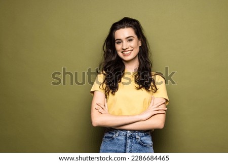 Young smiling attractive girl is posing in close pose with arms crossing on chest on khaki background looking at camera. Shy student, modest woman.