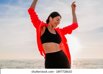 young smiling attractive slim woman doing sports in morning sunrise dancing on sea beach in sports wear, healthy lifestyle, listening to music on earphones, wearing pink windbreaker jacket, having fun