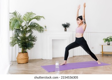 Young smiling attractive sporty Asian woman practicing yoga, doing Virabhadrasana 1 exercise, meditating in Warrior One yoga pose, indoor working out at home, wearing sportswear. Full length photo.
