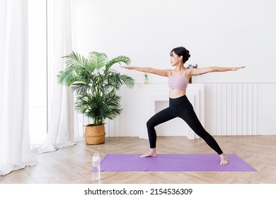 Young smiling attractive sporty Asian woman practicing yoga, doing Virabhadrasana 2 exercise, meditating in Warrior Two yoga pose, indoor working out at home, wearing sportswear. Full length photo.