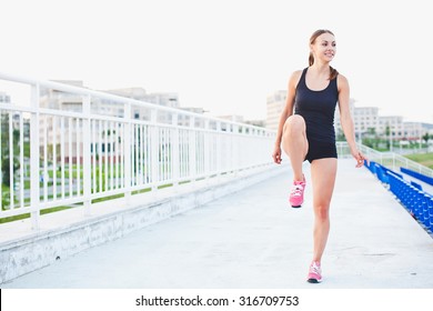 Young smiling attractive sporty girl runner in shorts and sleeveless shirt keeps her leg raised while warming up and stretching outdoors at stadium of university campus seen on background