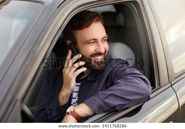 Young smiling attractive successful bearded man\
in a blue jacket and striped t-shirt, sits behind the wheel of the\
car in traffic, so as not to be bored called his friend and tells\
funny stories.