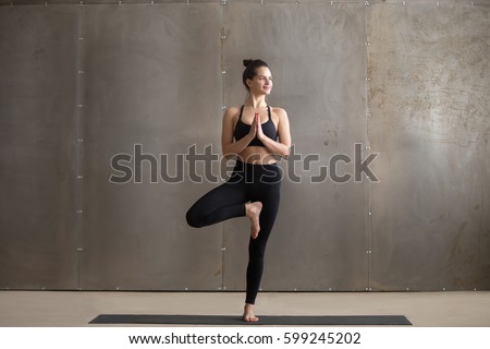 Young smiling attractive yogi woman practicing yoga, standing in Vrksasana exercise, Tree pose, working out, wearing black sportswear, cool urban style, full length, grey studio background 