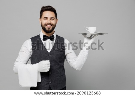 Young smiling barista male waiter butler man wear white shirt vest elegant uniform work at cafe hold plate coffee cup isolated on plain grey background studio portrait. Restaurant employee concept.