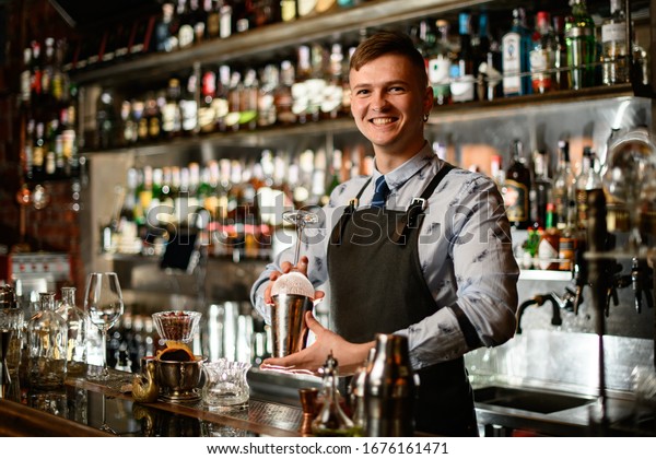 young\
smiling bartender in black apron preparing to make cocktail.\
Shelves with bottles of alcohol in\
background.