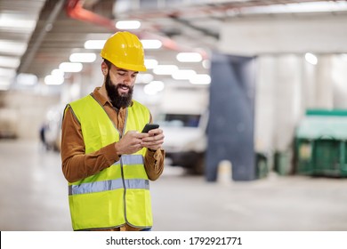Young smiling bearded construction worker standing in underground garage in construction process and using smart phone for texting. It’s break time.