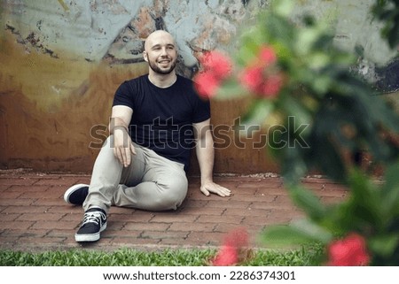 Young smiling bearded man sitting on the pavement in a park against a grungy wall. One happy masculine guy dressed casually relaxing on a street sidewalk