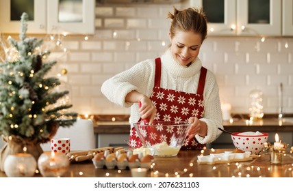Young smiling beautiful woman in apron making dough for christmas gingerbread cookies while cooking in cozy decorated kitchen, happy female cooking during winter holidays, making xmas sweets