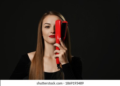 Young smiling beautiful woman with long silky straight hair in black clothes holding red hair straightener in hand over one eye over dark background. Haircare, beauty, wellness, styling concept