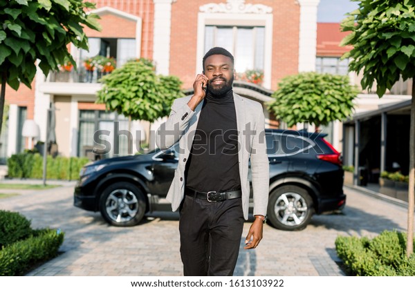 Young smiling black business man in a suit\
talking by smartphone walking outdoors near a black car and modern\
brick buildings