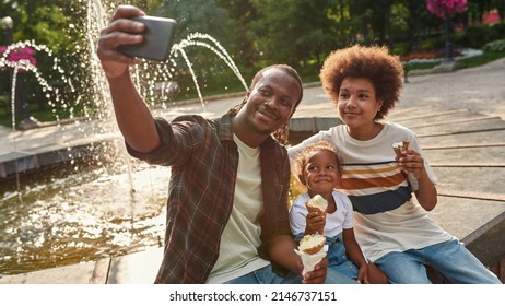 Young Smiling Black Family With Ice Cream Cones Taking Selfie On Smartphone Near Fountain In City Park. Family Relationship And Spending Time Together. Fatherhood And Parenting. Sunny Summer Day