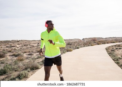 Young Smiling Black Runner In A Yellow Rain Jacket Who Enjoys Listening To Music On His Cell Phone While Training.