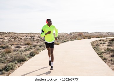 Young Smiling Black Runner In A Yellow Rain Jacket Who Enjoys Listening To Music On His Cell Phone While Training.