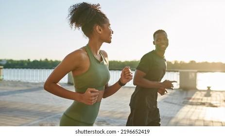 Young smiling black sports couple running on river promenade in city. Attractive girl and athletic man wearing sportswear and looking at each other. Concept of modern healthy lifestyle. Warm sunny day - Powered by Shutterstock