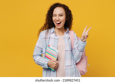 Young smiling black teen girl student she wear casual clothes backpack bag hold books showing victory sign blink eye isolated on plain yellow color background. High school university college concept - Powered by Shutterstock