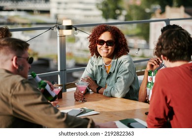 Young Smiling Black Woman With Pink Cocktail In Martini Glass Sitting By Table In Front Of Her Friends Enjoying Gathering In Outdoor Cafe