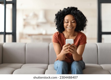 Young Smiling Black Woman With Smartphone Resting On Couch At Home, Happy African American Female Sitting On Sofa And Using Mobile Phone, Messaging With Friends Or Browsing New App For Shopping - Powered by Shutterstock