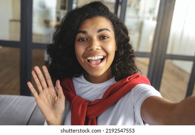 Young Smiling Black Woman Using Camera Of Mobile Phone Taking Selfie Smiling At The Camera In The Living Room At Home. Showing Hello Gesture, Smartphone Webcam