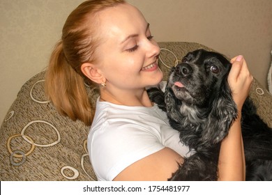 Young Smiling Blond Caucasian Woman In White T-shirt Seats On Chair And Hug Black Russian Spaniel At Home. Happy Girl Holding Cute Black Dog Looking Right Into The Camera. Owner And Pet Love