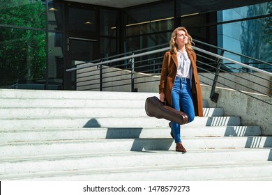 Young smiling blond female student musician in brown coat with violin in leather case going down the stairs after conservatory class or concert at sunny day. Music, education and arts concept