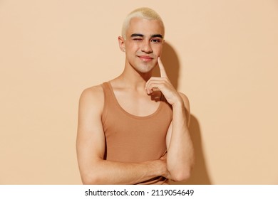 Young smiling blond latin gay man with make up in beige tank shirt hug himself look camera blink prop up face isolated on plain light ocher background studio portrait People lgbt lifestyle concept.