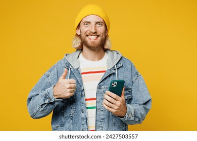 Young smiling blond man he wearing denim shirt hoody beanie hat casual clothes hold in hand use mobile cell phone show thumb up isolated on plain yellow background studio portrait. Lifestyle concept - Powered by Shutterstock