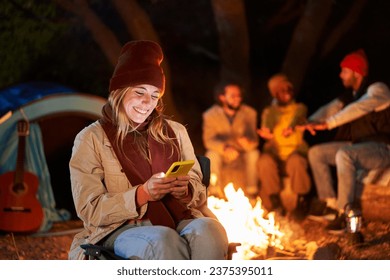 Young smiling blonde girl using cell phone at campsite at winter night. Cheerful millennial woman sitting by campfire looking at mobile with friends in background. People addicted to social networks.  - Powered by Shutterstock