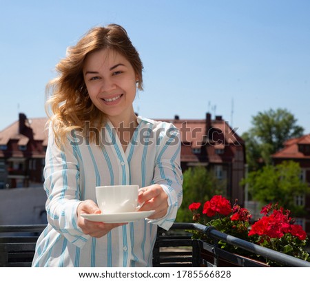 young smiling blonde holds out a white cup in front of her offering a drink. Against the background of the blue summer sky