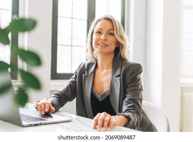 Young smiling blonde woman with long hair in stylish grey suit working at laptop in the bright modern office - Powered by Shutterstock