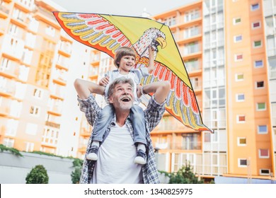 Young Smiling Boy Playing With Colorful Kite Outside Sitting On Grandfather's Shoulders. Little Grandson With His Grandfather Playing With A Kite On The Urban Background.