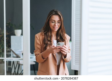 Young smiling brunette business woman in glasses with long hair in stylish beige suit using smartphone in modern office - Powered by Shutterstock