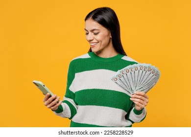 Young smiling brunette latin woman wear casual cozy green knitted sweater use mobile cell phone hold in hand fan of cash money in dollar banknotes isolated on plain yellow background studio portrait - Shutterstock ID 2258228529