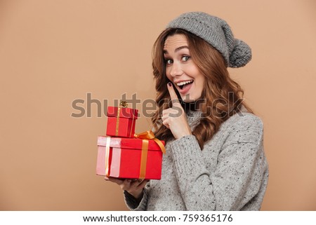 Young smiling brunette woman showing silence gesture while holding gift boxes, looking at camera, isolated on beige background