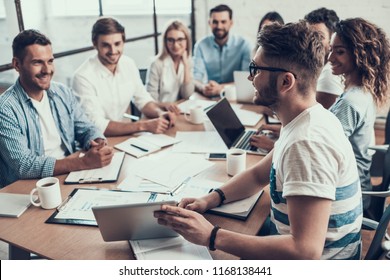 Young Smiling Business People on Meeting in Office. Man showing Project on Tablet to Group of Young Happy Collegues Sitting Together at Table Working on Laptops in Modern Office. Teamwork Concept