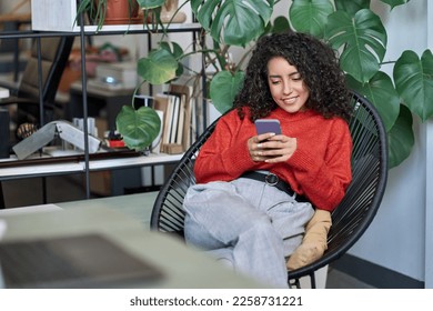 Young smiling business woman looking at smartphone using cellphone mobile cellular, professional female worker or student typing on mobile cell phone sitting in chair with green plants around. - Powered by Shutterstock