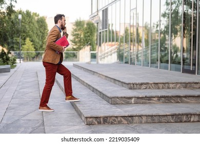 Young Smiling Businessman Carrying Documents And Calling While Moving Forward. Man In Casual Clothes Walking Near Modern Office Building, Full Length, Copy Space. Business And Success Concept