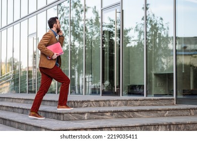 Young Smiling Businessman Carrying Documents And Calling While Moving Forward. Man In Casual Clothes Walking Near Modern Office Building, Full Length, Copy Space. Business And Success Concept