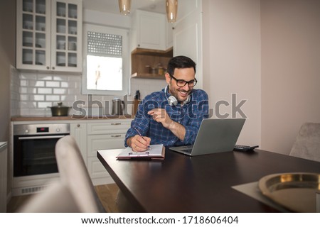 Young smiling businessman working from home writing notes in notebook while using his laptop and wearing headphones around his neck.