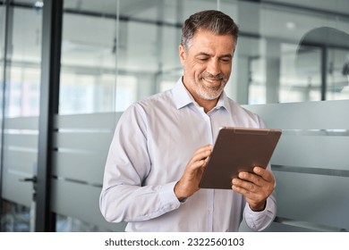 Young smiling busy mature male manager using tablet standing in office. Older mid aged professional business man company executive holding tab digital computer managing financial banking data. - Powered by Shutterstock