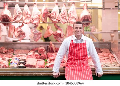 A young smiling butcher in a red apron standing and leaning his back against the butcher shop window.