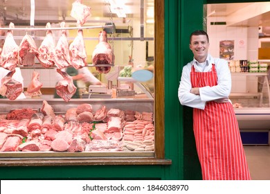 A Young Smiling Butcher In Red Apron Leaning Against Butcher Shop Doorway Next To The Display Window.