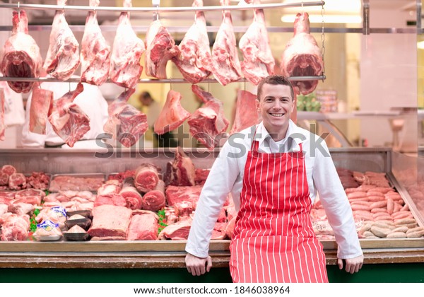 A young smiling butcher\
in a red apron standing and leaning his back against the butcher\
shop window.